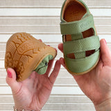 Close up of adult holding Froddo's olive barefoot kids sandals showing rubber sole detail in one hand and velcro strap detail on other hand