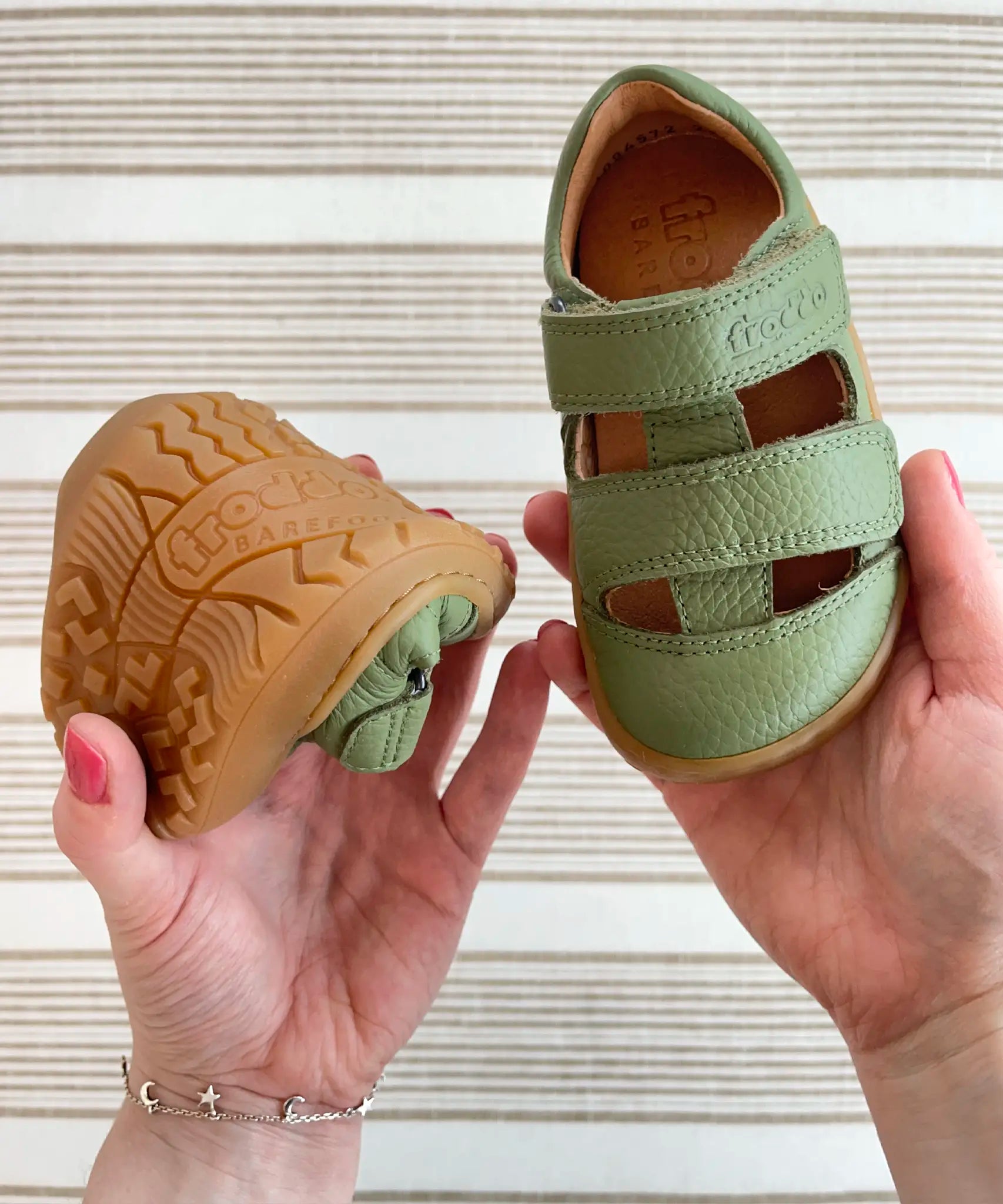 Close up of adult holding Froddo's olive barefoot kids sandals showing rubber sole detail in one hand and velcro strap detail on other hand