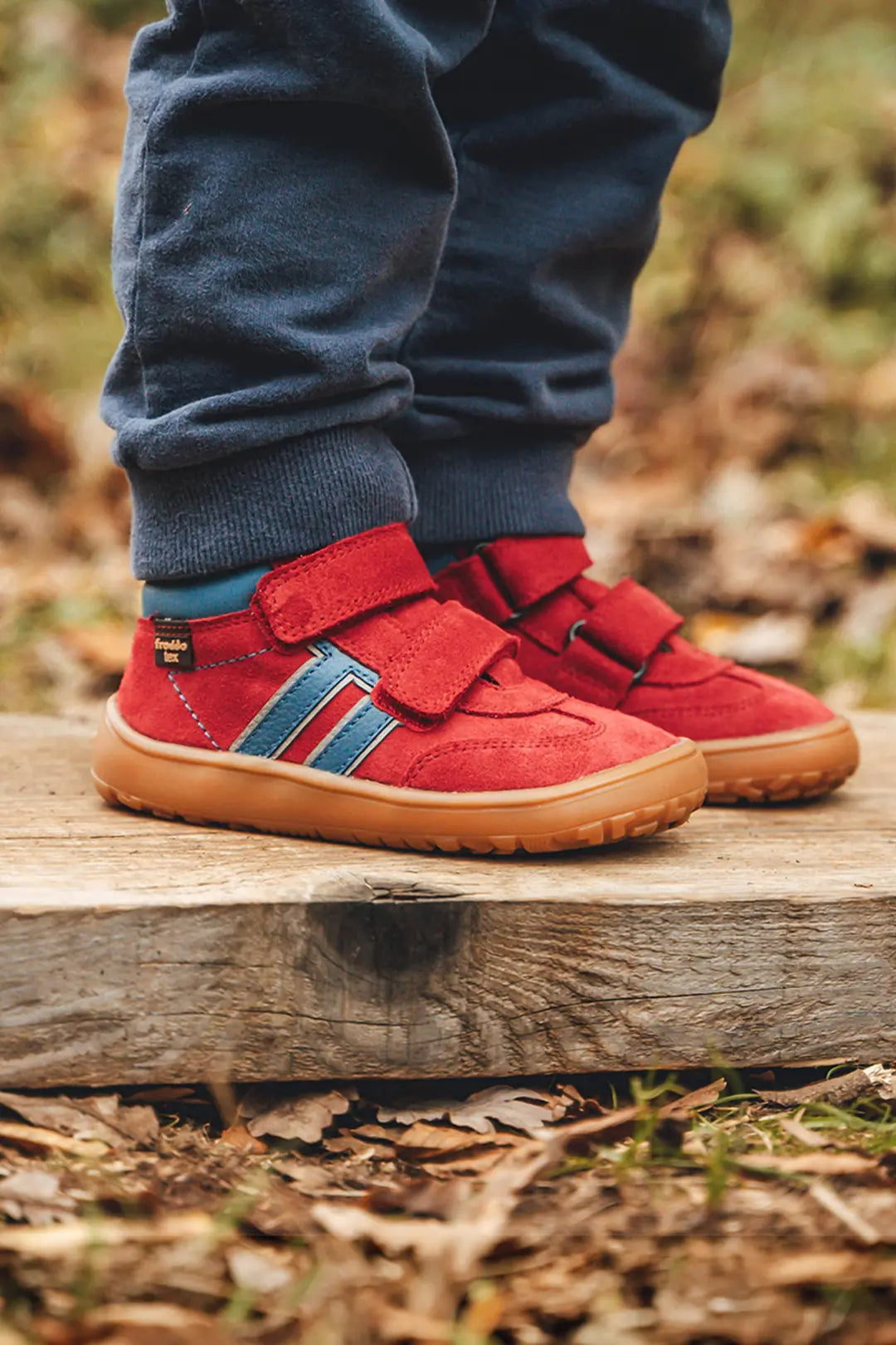 Red toddler Froddo Barefoot Trainers with blue and red side stripes and tan soles, shown on a child standing on a wooden log outdoors.