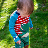 A child standing outside and wearing the Frugi kids alfie raglan top in berry red/ breton frog. A GOTS organic cotton top with long sleeves with a playful bee applique on the wrist and a green frog on the front.