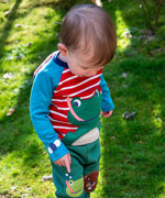 A child standing outside and wearing the Frugi kids alfie raglan top in berry red/ breton frog. A GOTS organic cotton top with long sleeves with a playful bee applique on the wrist and a green frog on the front.