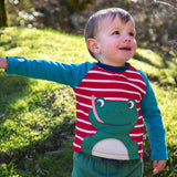 A child wearing the Frugi Alfie raglan kids top in red breton frog available at Babipur. A GOTS organic cotton top with blue long sleeves and a playful green frog applique on the red and white stripe front panel.
