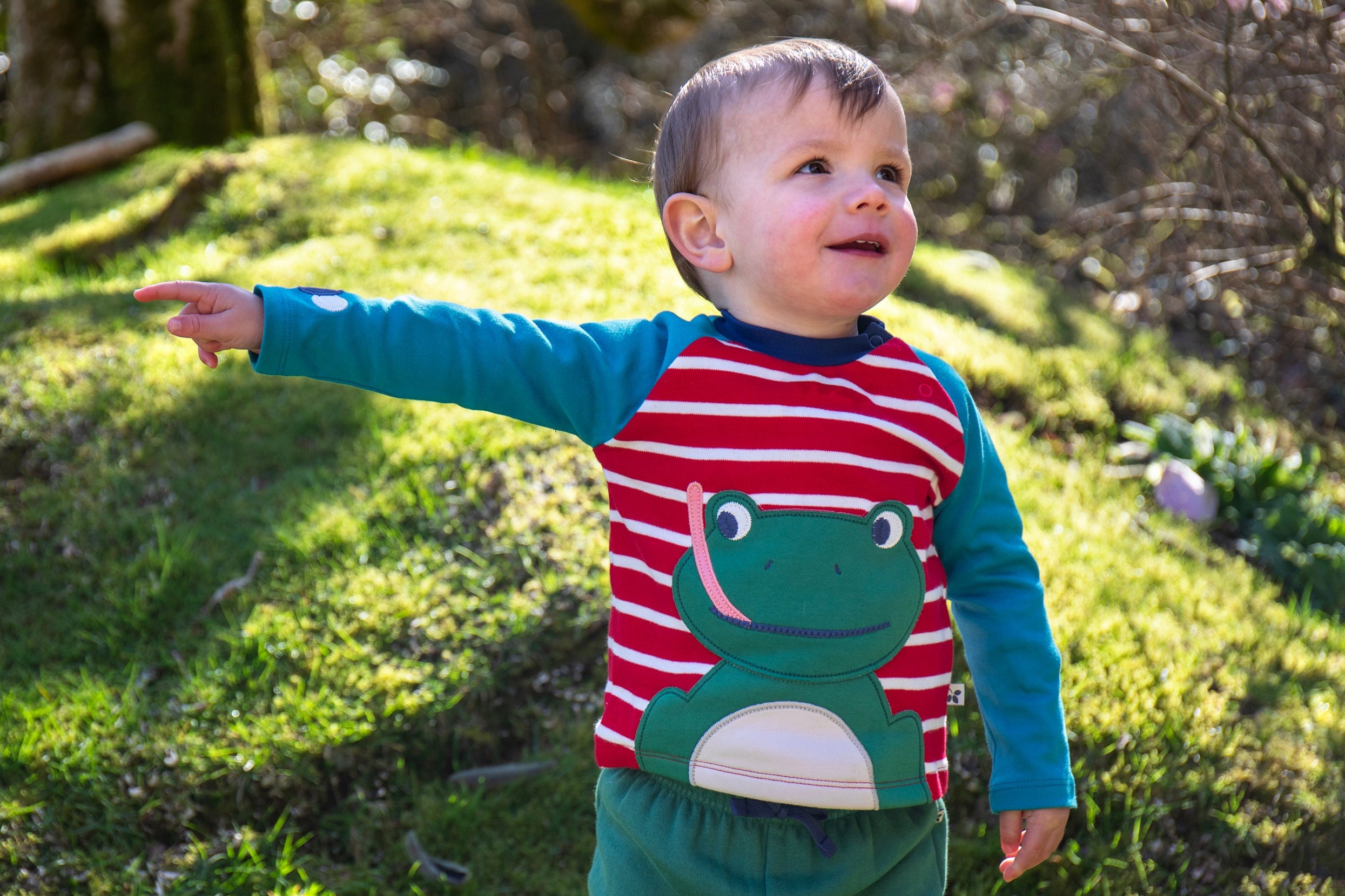 A child wearing the Frugi Alfie raglan kids top in red breton frog available at Babipur. A GOTS organic cotton top with blue long sleeves and a playful green frog applique on the red and white stripe front panel.
