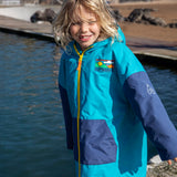 Child wearing Frugi's blue Atlantic hooded changing robe with navy sleeves shows a full length zip with water and the beach in the background