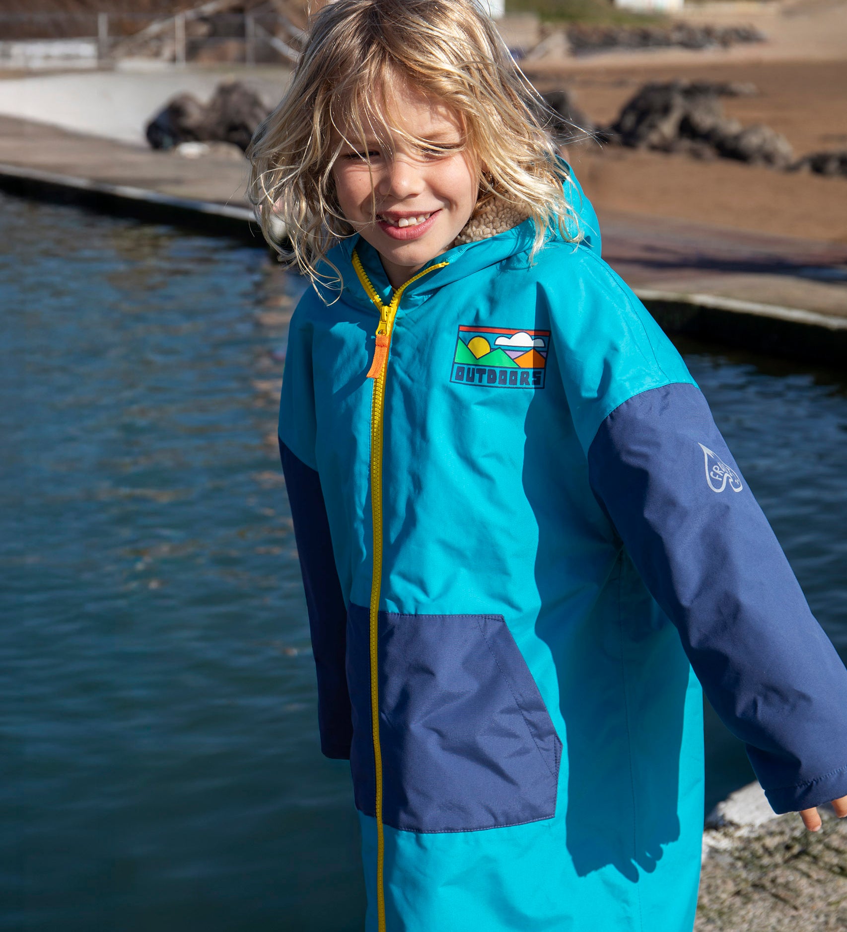 Child wearing Frugi's blue Atlantic hooded changing robe with navy sleeves shows a full length zip with water and the beach in the background