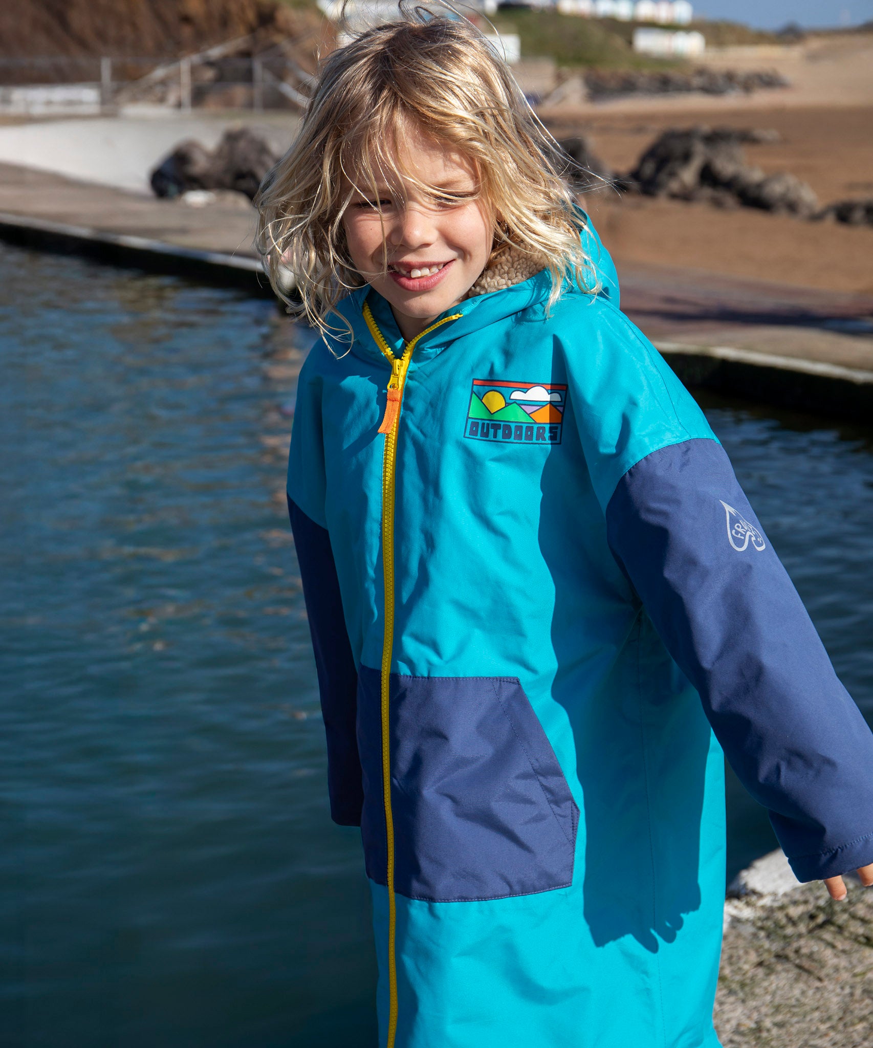 Child wearing Frugi's blue Atlantic hooded changing robe with navy sleeves shows a full length zip with water and the beach in the background