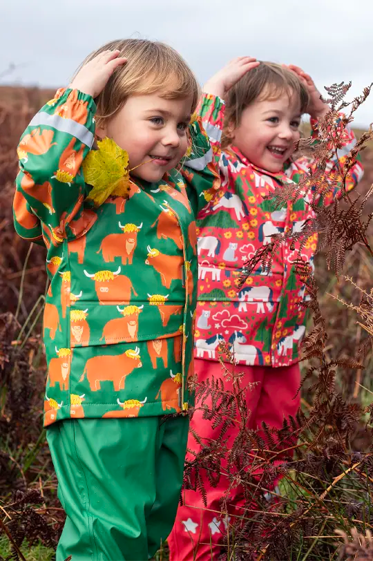Frugi Puddle Buster Coat in green Highland Grazing all-over print with elasticated sleeves and reflective details, worn by child playing outdoors.