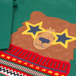 A closer look at the bear applique on the Frugi Terrific Applique Top - Teal/Fairisle Bear. The bear is light brown, with yellow star sunglasses, and wears a red patterned top, on teal green fabric