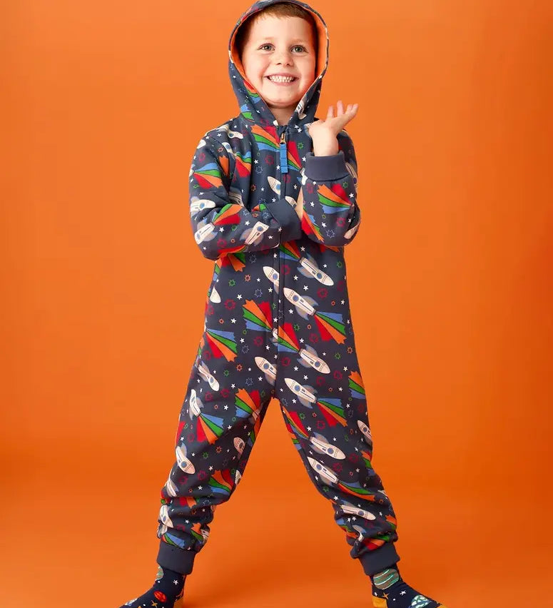 Frugi big organic cotton snuggle suit in blue with rainbow rocket and planet design, worn by child with hood up.