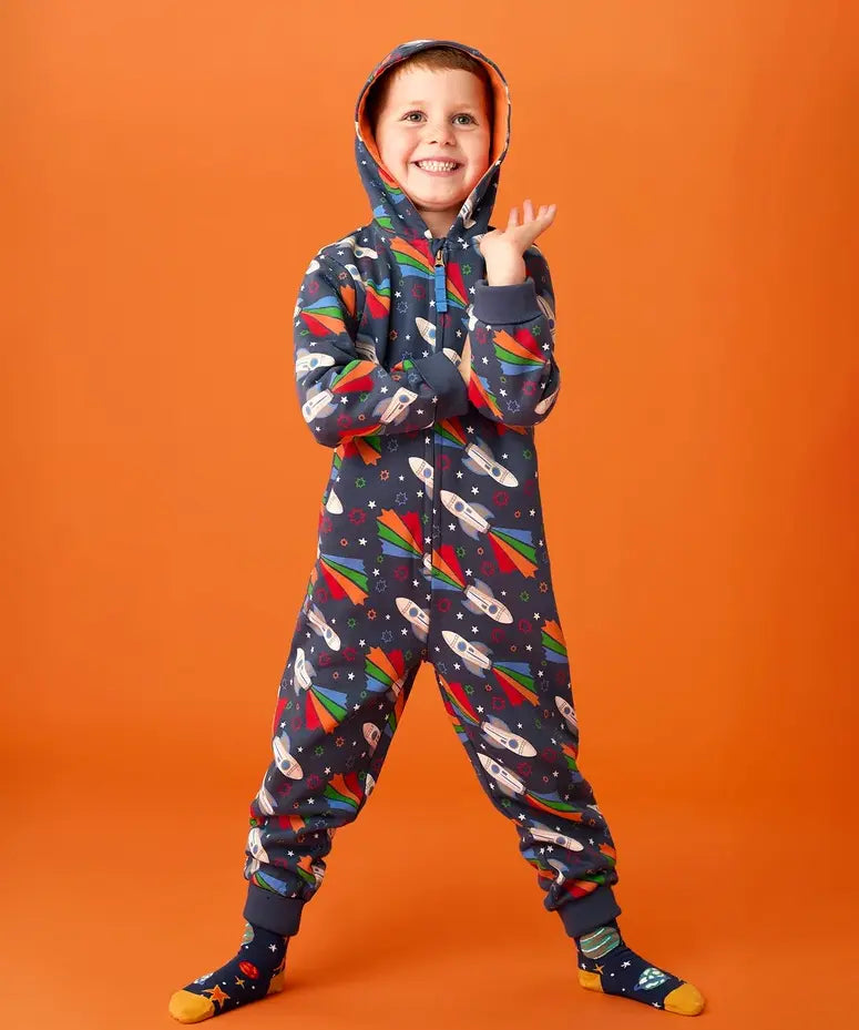 Frugi big organic cotton snuggle suit in blue with rainbow rocket and planet design, worn by child with hood up.