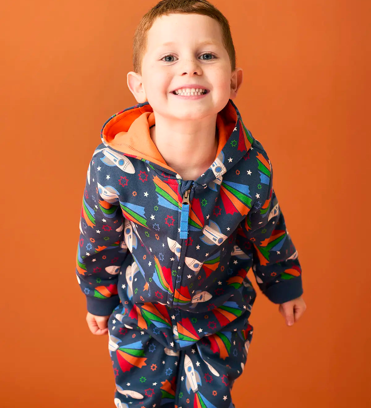 Child wearing Frugi's GOTS organic cotton big hooded snuggle suit in navy with repeating flying rocket prints and a orange inner liner 