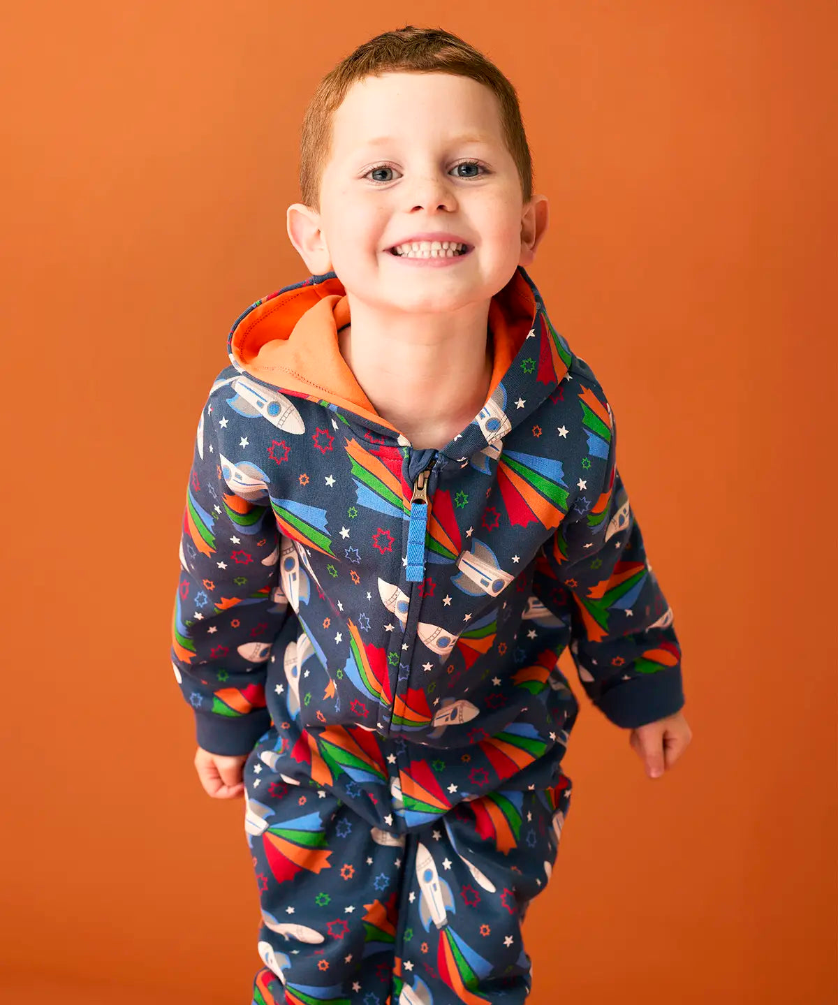 Child wearing Frugi's GOTS organic cotton big hooded snuggle suit in navy with repeating flying rocket prints and a orange inner liner 