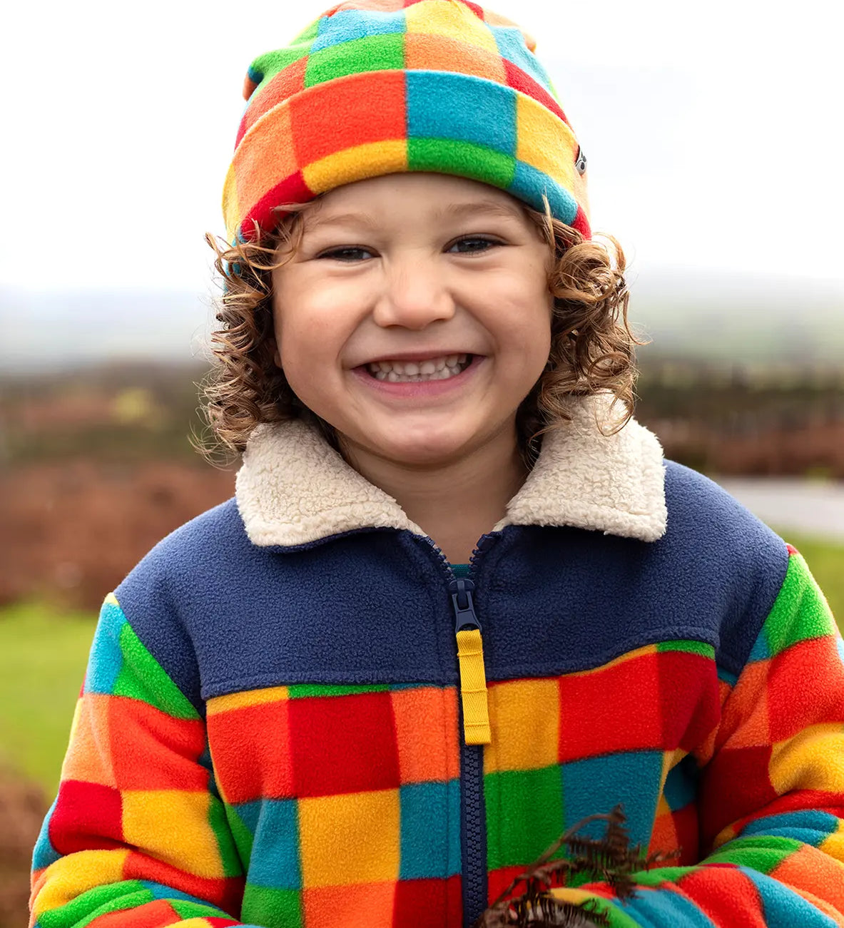Frugi Blazey Reversible Fleece Jacket in Kaleidoscope rainbow checkered print, worn by child with matching fleece hat.