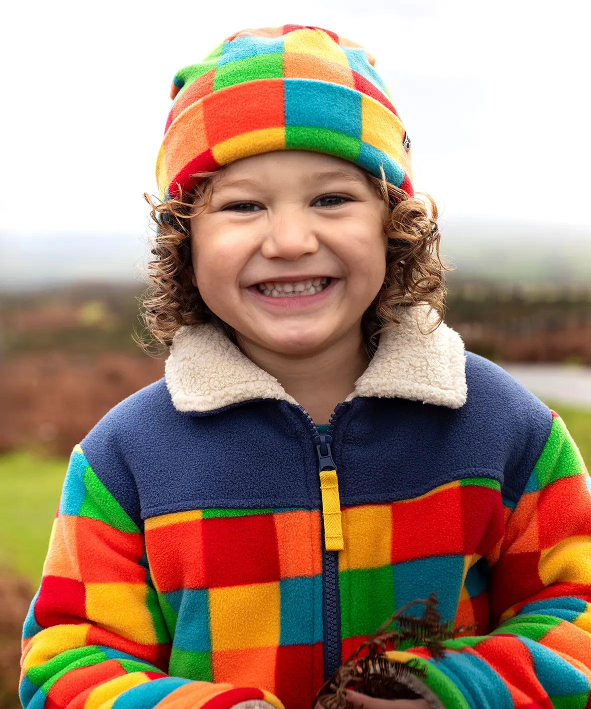 Frugi Blazey Reversible Fleece Jacket in Kaleidoscope rainbow checkered print, worn by child with matching fleece hat.