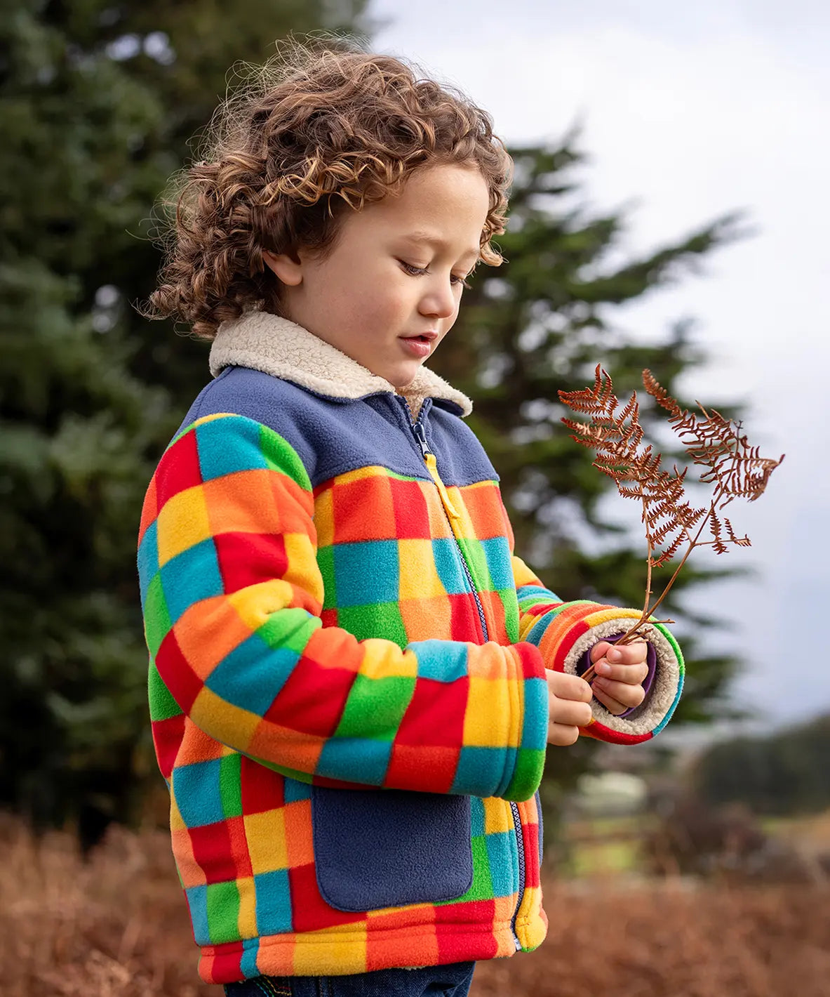 Frugi Blazey Reversible Fleece Jacket with cream collar in Kaleidoscope rainbow checkered print, worn by child with curly hair.