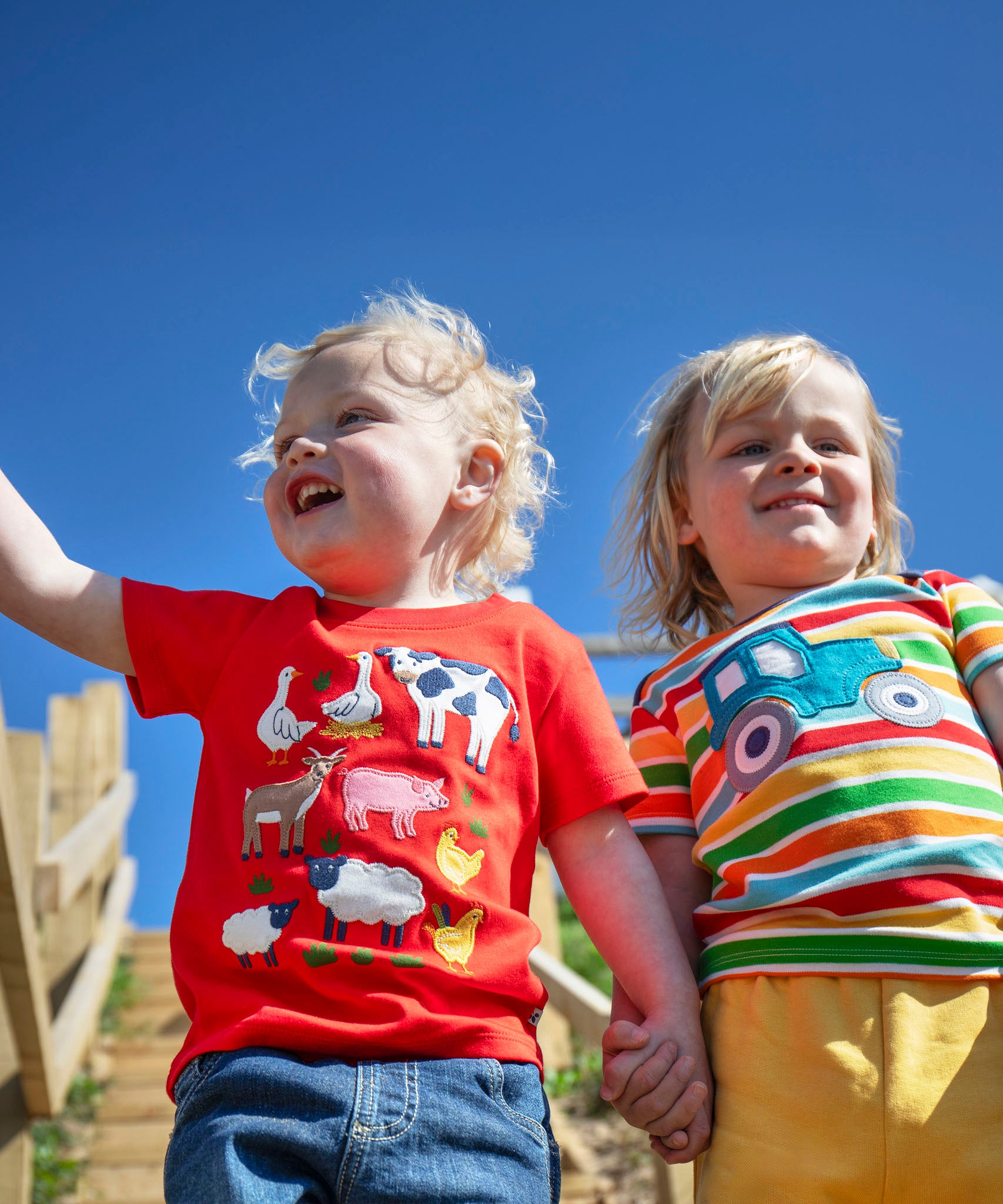 Child wearing Frugi's happy tractor bobster applique t-shirt outdoors next to a child wearing a red tshirt with farm animals