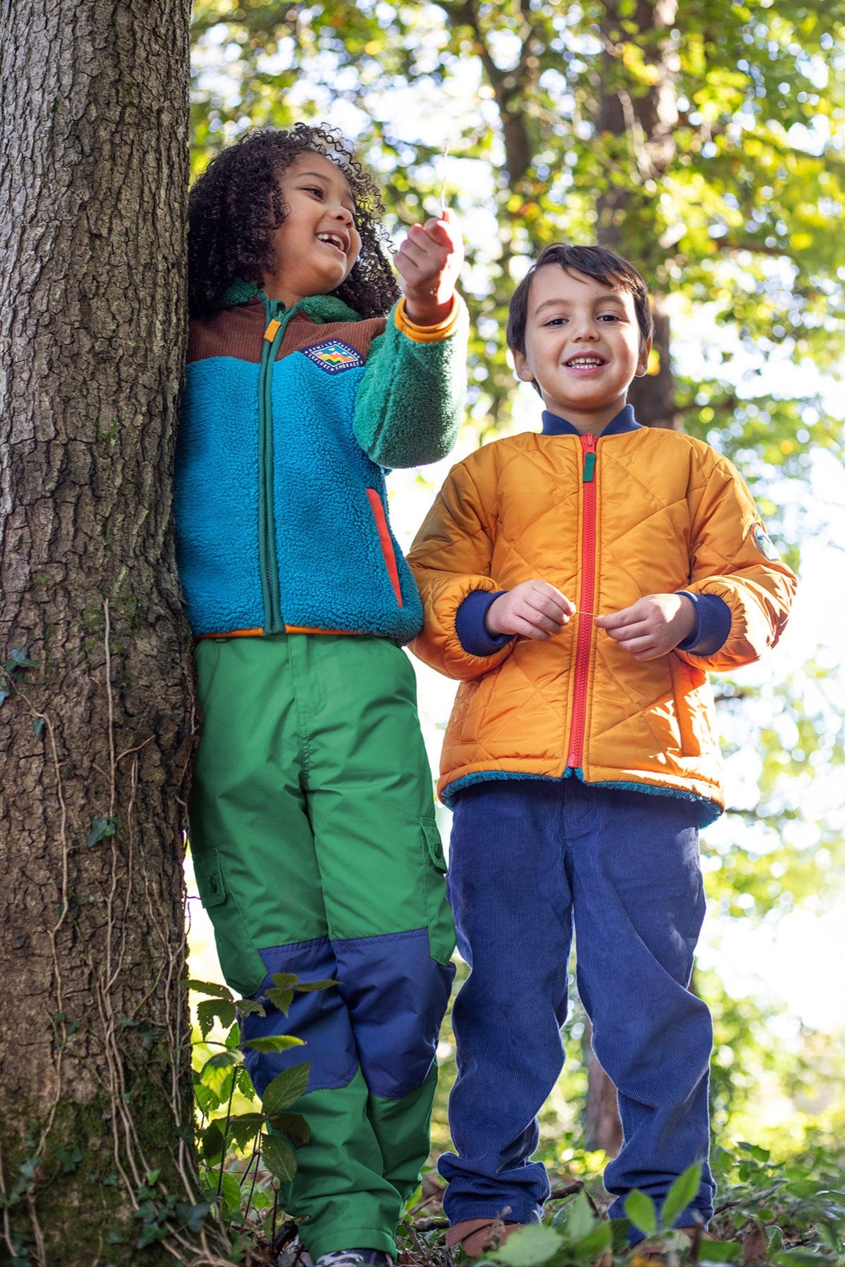 A child wearing the Frugi kids braken fleece cord jacket in holly green/hotchpotch and standing next to a child wearing the Frugi robin reversible quilted jacket - gold / blue