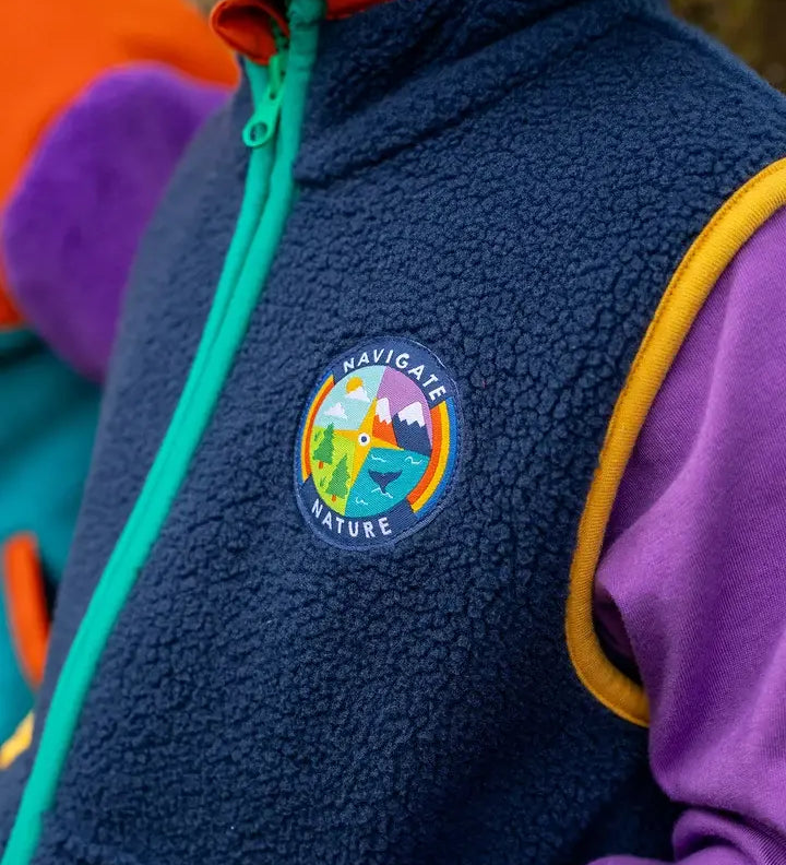 Frugi navy borg fleece gilet with blue and orange piping and 'navigate nature' badge, worn by child with purple hoodie.