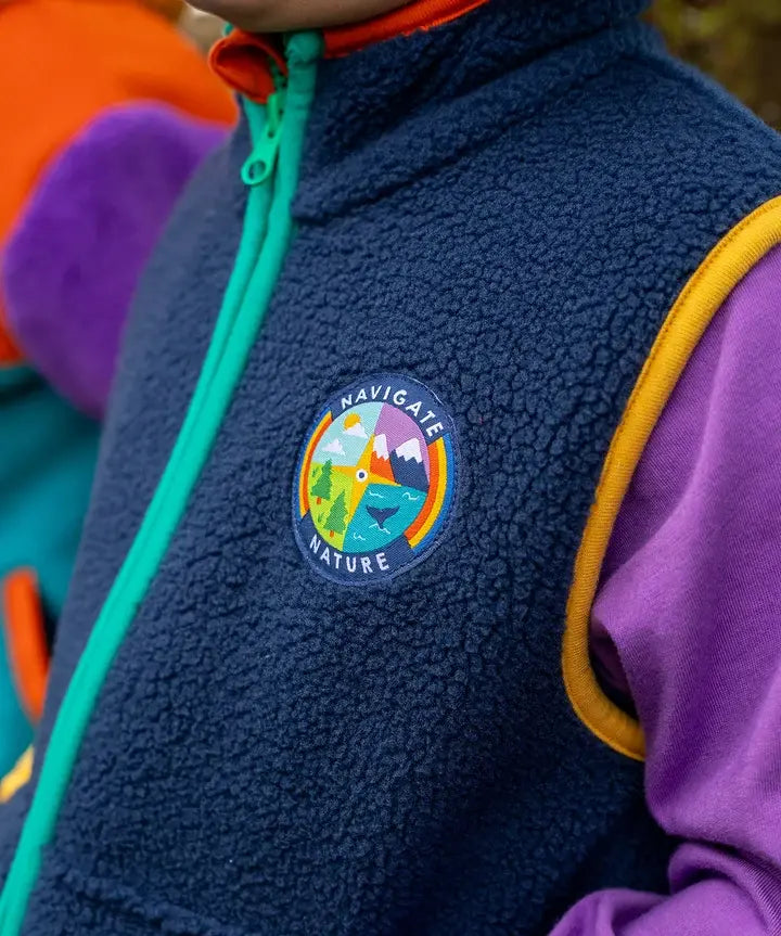 Frugi navy borg fleece gilet with blue and orange piping and 'navigate nature' badge, worn by child with purple hoodie.