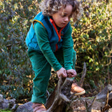 A child playing on a fallen tree trunk and wearing the Frugi bude borg fleece kids gilet in boat blue available at Babipur. A thick blue fleece with orange trim. 