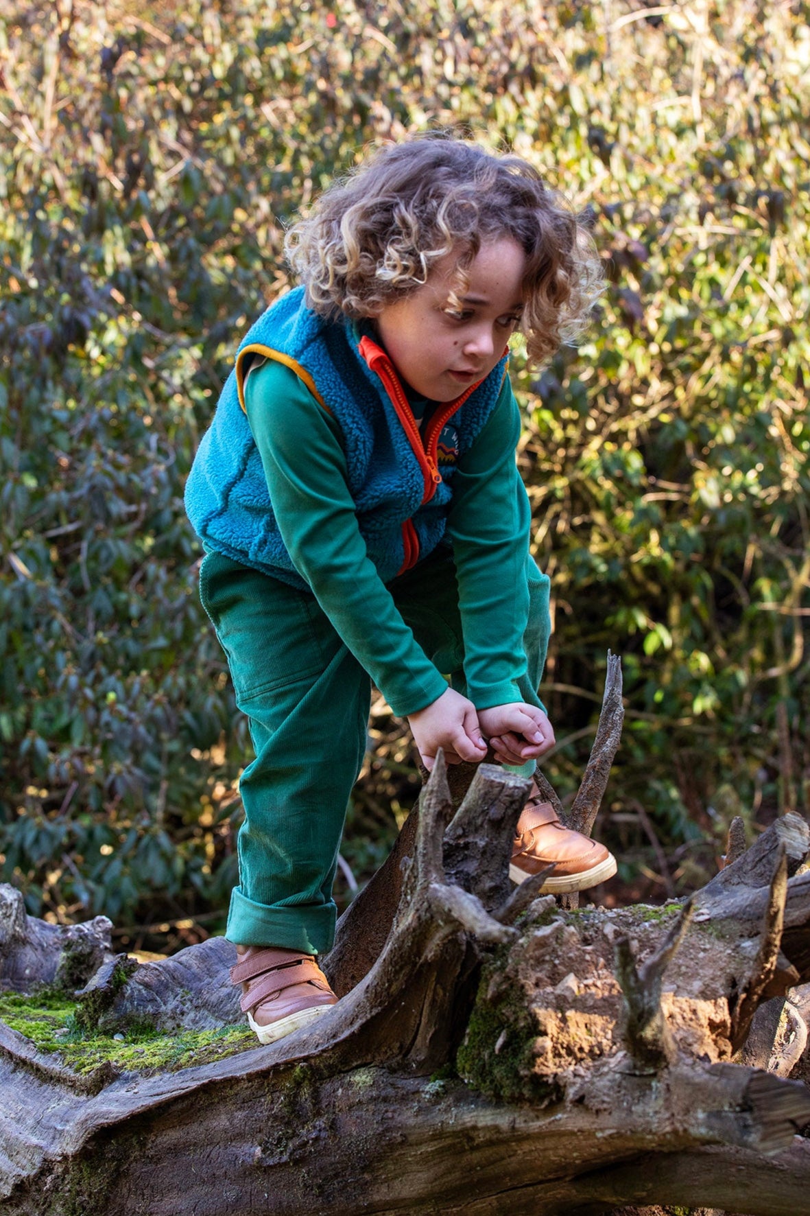A child playing on a fallen tree trunk and wearing the Frugi bude borg fleece kids gilet in boat blue available at Babipur. A thick blue fleece with orange trim. 