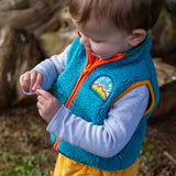 A toddler playing and wearing the Frugi bude borg fleece kids gilet in boat blue available at Babipur. A thick blue fleece with orange trim and a keep it wild slogan patch on the chest. Features a full zip fastener 
