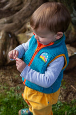 A toddler playing and wearing the Frugi bude borg fleece kids gilet in boat blue available at Babipur. A thick blue fleece with orange trim and a keep it wild slogan patch on the chest. Features a full zip fastener 