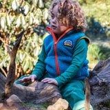 A child standing on a fallen tree trunk and wearing the Frugi bude borg fleece kids gilet in boat blue available at Babipur. A thick blue fleece with orange trim. Features a keep it wild mountain patch