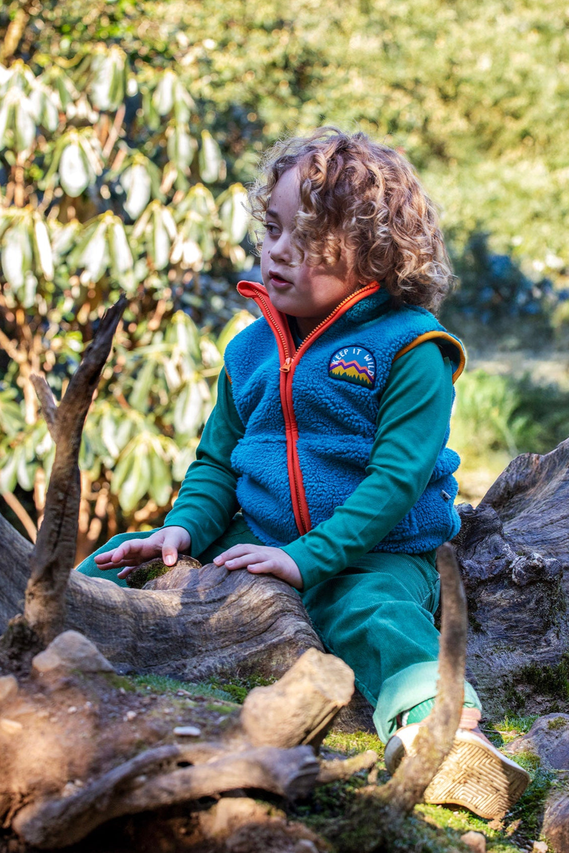 A child standing on a fallen tree trunk and wearing the Frugi bude borg fleece kids gilet in boat blue available at Babipur. A thick blue fleece with orange trim. Features a keep it wild mountain patch