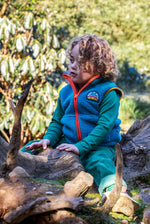 A child standing on a fallen tree trunk and wearing the Frugi bude borg fleece kids gilet in boat blue available at Babipur. A thick blue fleece with orange trim. Features a keep it wild mountain patch