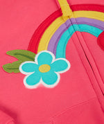 A close up of the light pink zipped hoodie laid flat. Showing the detailing on the rainbow patch and blue flower running across the middle.
