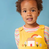 Frugi Catrin reversible yellow cord playsuit with house applique paired with a striped long sleeve top, worn by little girl.
