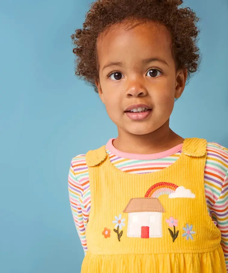 Frugi Catrin reversible yellow cord playsuit with house applique paired with a striped long sleeve top, worn by little girl.