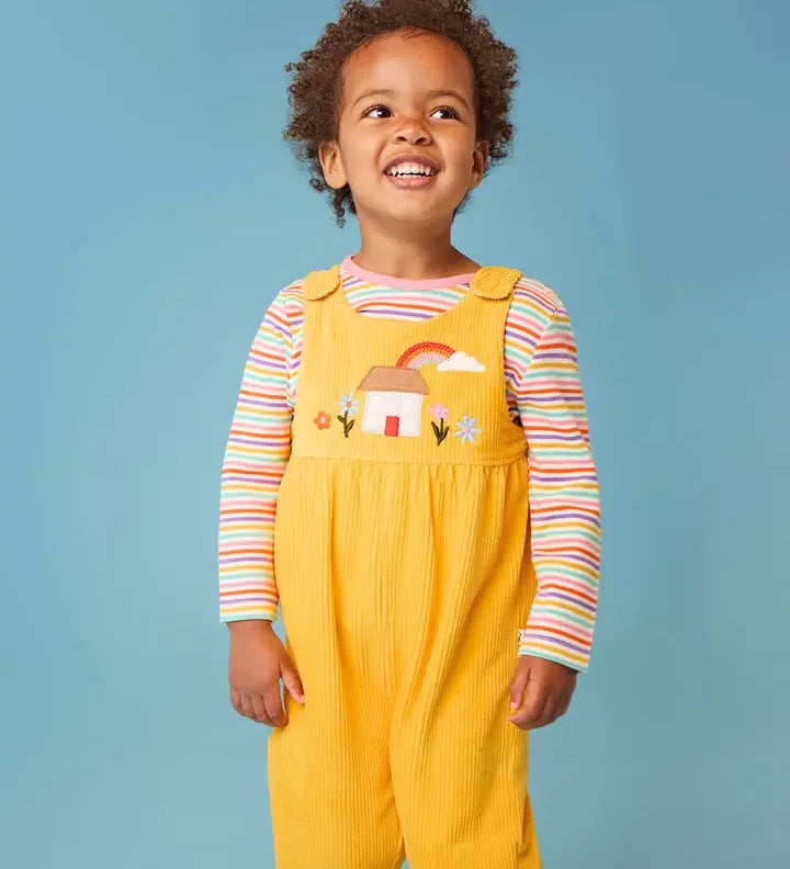Frugi Catrin reversible dungarees in yellow cord with house applique on bib paired with a striped long sleeve top, worn by little girl.