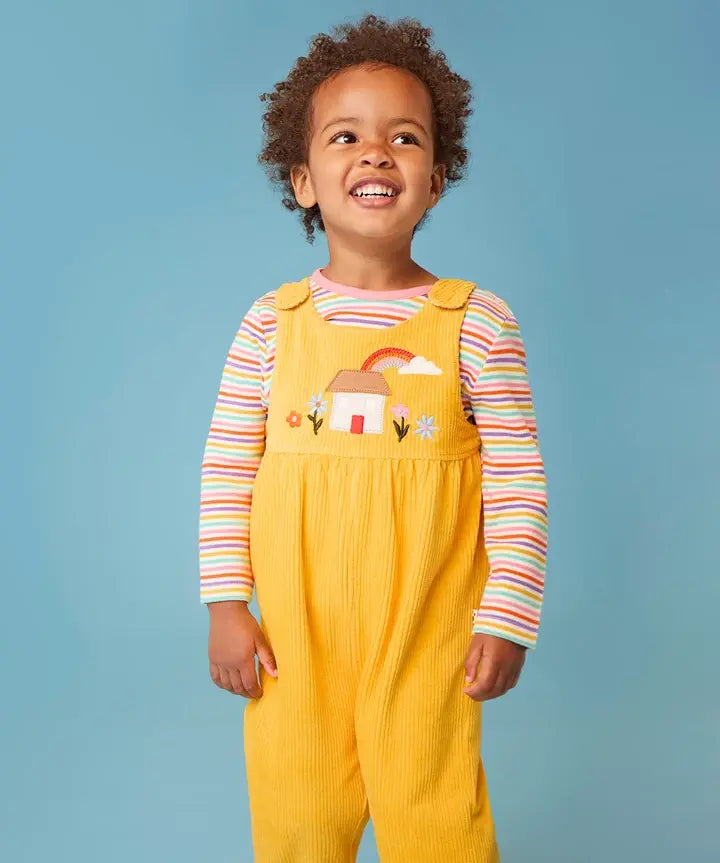 Frugi Catrin reversible dungarees in yellow cord with house applique on bib paired with a striped long sleeve top, worn by little girl.