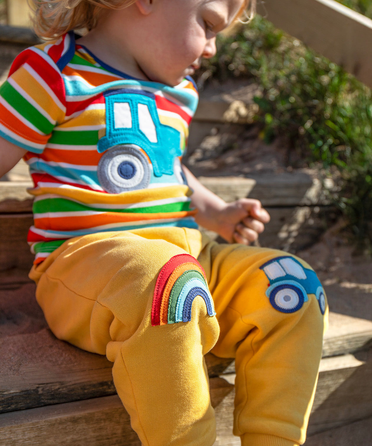 A smiling child sitting outside on wooden stairs. Wearing a multicoloured stripe t-shirt and bright yellow joggers with a rainbow applique on one knee and a blue tractor on the other knee