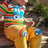 A smiling child sitting outside on wooden stairs. Wearing a multicoloured stripe t-shirt and bright yellow joggers with a rainbow applique on one knee and a blue tractor on the other knee