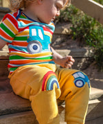 A smiling child sitting outside on wooden stairs. Wearing a multicoloured stripe t-shirt and bright yellow joggers with a rainbow applique on one knee and a blue tractor on the other knee
