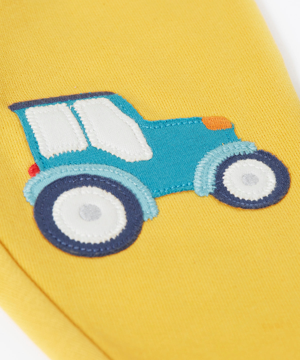 A close up of the bright yellow Frugi joggers laid flat showing the detailing on the blue tractor applique