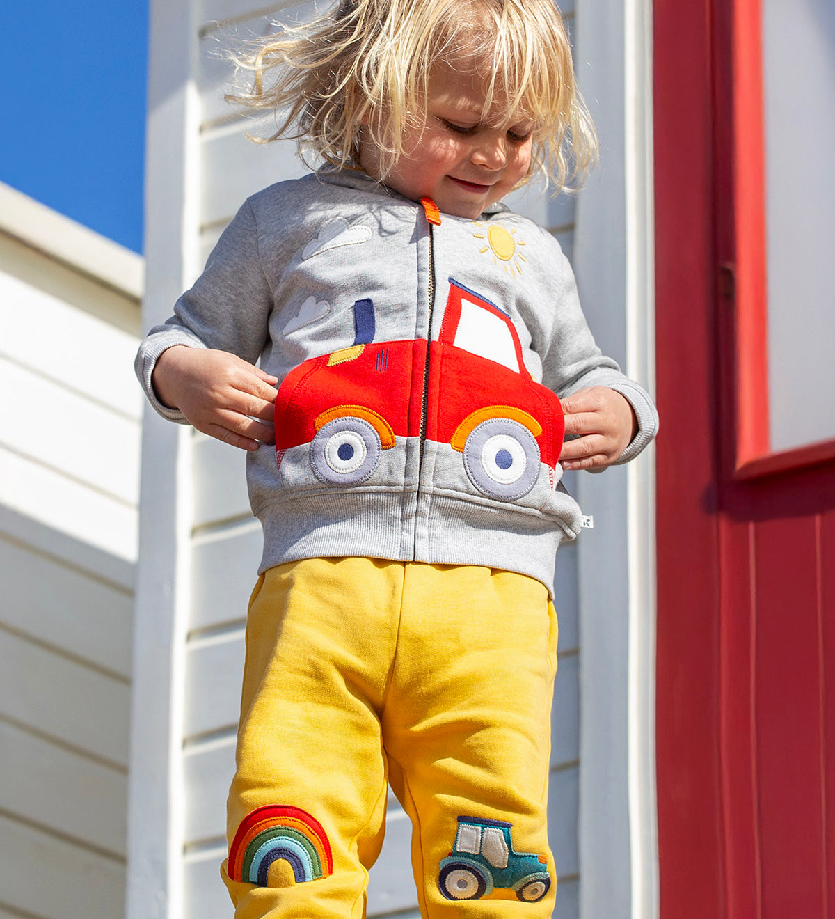 A smiling child standing outside in front of a white beach hut with a red door. Wearing a grey zip hoody and the bright yellow joggers