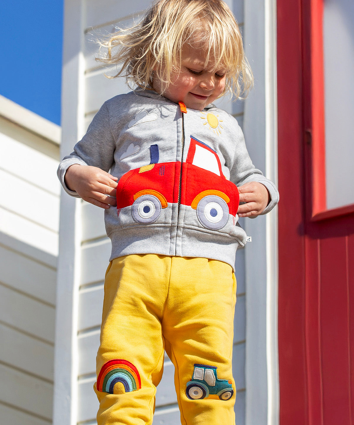 A smiling child standing outside in front of a white beach hut with a red door. Wearing a grey zip hoody and the bright yellow joggers