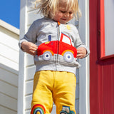 A smiling child standing outside in front of a white beach hut with a red door. Wearing a grey zip hoody and the bright yellow joggers