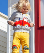 A smiling child standing outside in front of a white beach hut with a red door. Wearing a grey zip hoody and the bright yellow joggers
