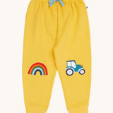 Frugi organic cotton joggers laid flat. In bright yellow with a rainbow appliqué on one knee and a blue tractor on the other knee. Features an elasticated waist band with blue tie