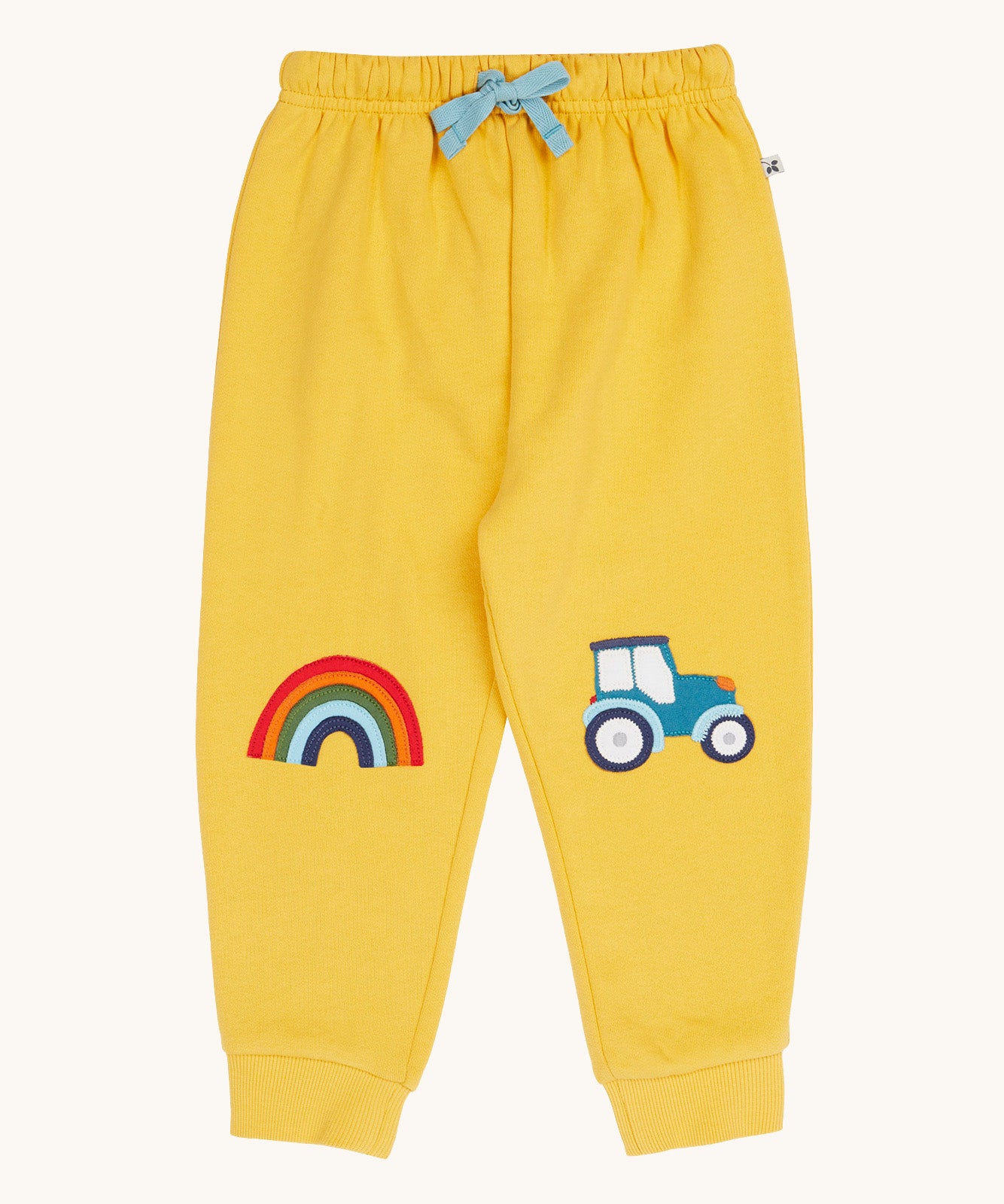 Frugi organic cotton joggers laid flat. In bright yellow with a rainbow appliqué on one knee and a blue tractor on the other knee. Features an elasticated waist band with blue tie