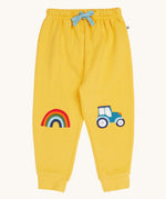 Frugi organic cotton joggers laid flat. In bright yellow with a rainbow appliqué on one knee and a blue tractor on the other knee. Features an elasticated waist band with blue tie