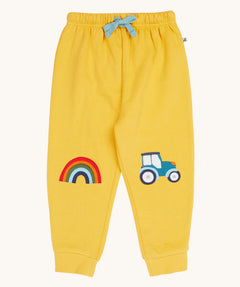 Frugi organic cotton joggers laid flat. In bright yellow with a rainbow appliqué on one knee and a blue tractor on the other knee. Features an elasticated waist band with blue tie
