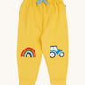 Frugi organic cotton joggers laid flat. In bright yellow with a rainbow appliqué on one knee and a blue tractor on the other knee. Features an elasticated waist band with blue tie