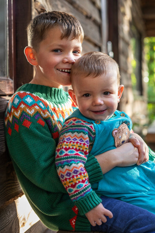 Two boys dressed in Christmas Frugi - one in a green knitted fairisle jumper, the other is wearing a pair of blue cord dungarees paired with a fairisle bear jumper, all made from GOTS organic cotton - to represent Frugi Autumn Winter at Babipur.
