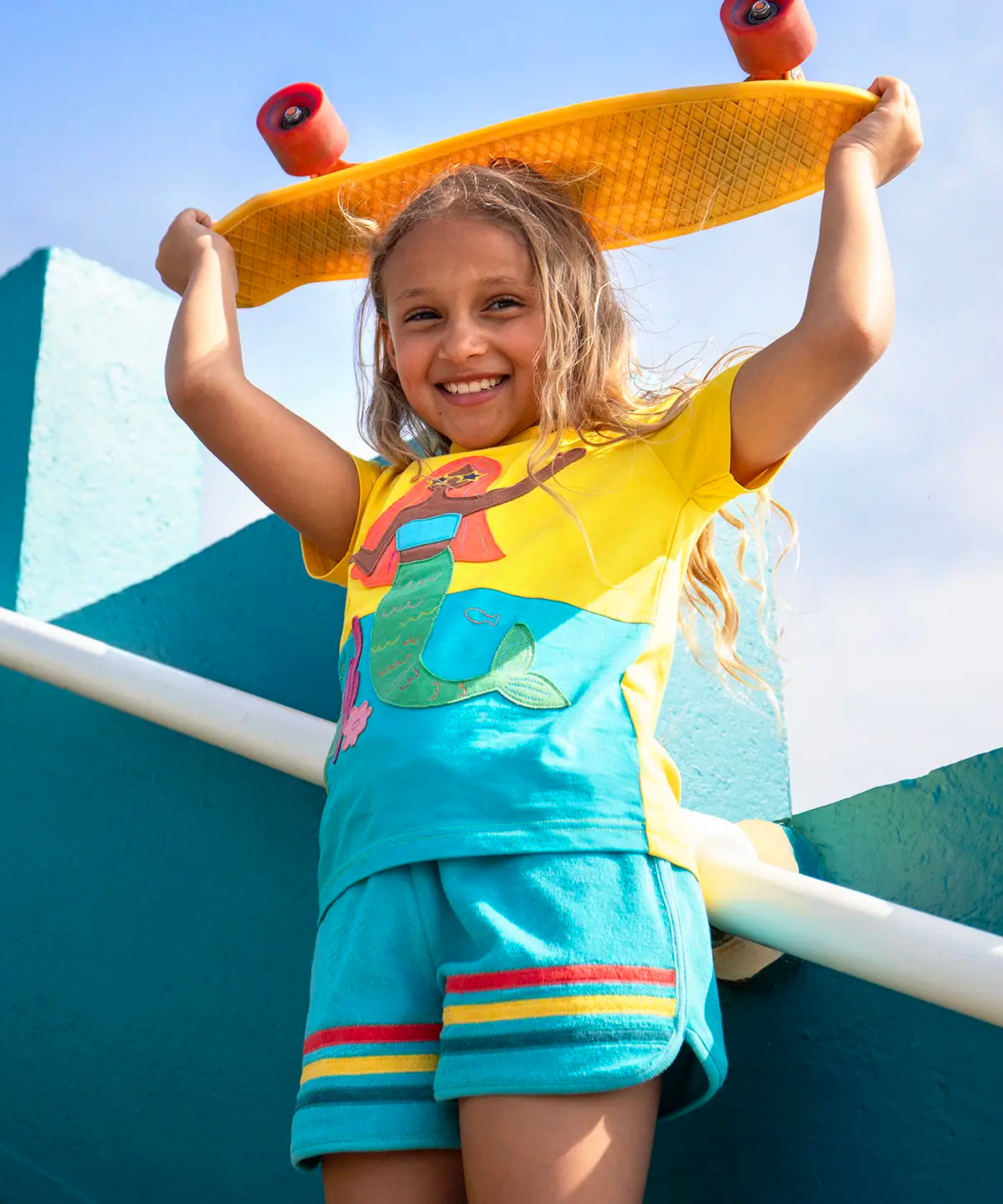 Child wearing Frugi's coast towelling shorts in light blue with red, yellow, blue stripe on hem and holding a skateboard